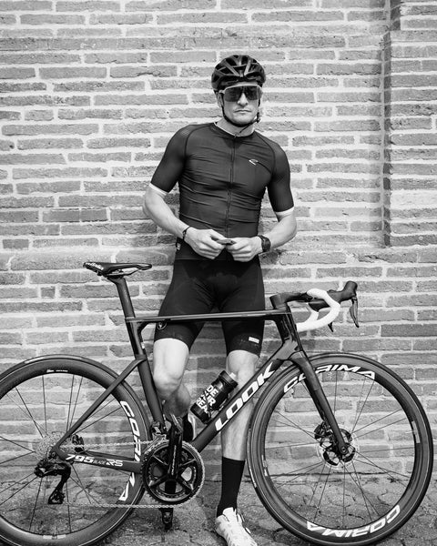 Cyclist in cycling gear standing next to a bicycle against a brick wall.